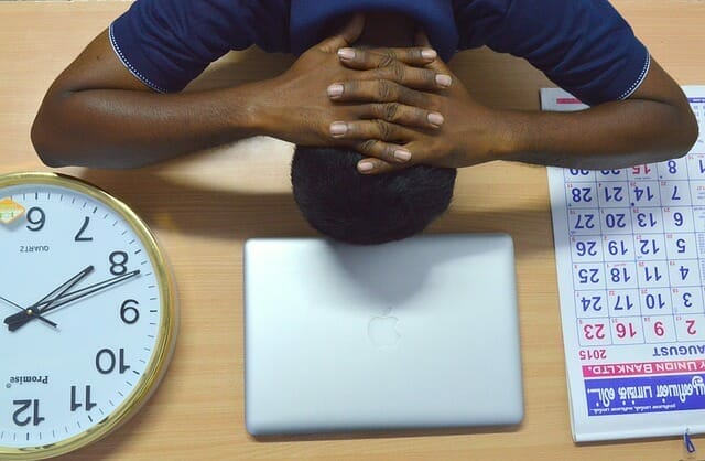  Um homem muito cansado mantém a cabeça na secretária, junto ao portátil, calendário e relógio.